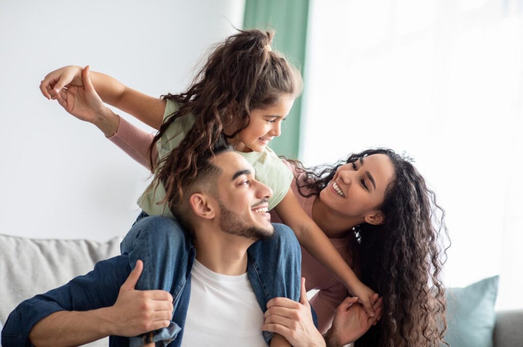 Portrait Of Cheerful Middle Eastern Family Of Three Having Fun Together At Home. Young Arabic Parents Playing With Their Little Daughter In Living Room, Mom, Dad And Child Smiling And Laughing.