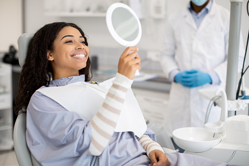 Patient in dental chair wearing a bib and holding a mirror, with dentist in mask and gloves nearby during check-up in a clinical setting.